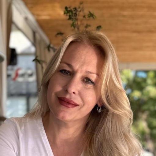 Selfie of a smiling blonde woman in a white top on an outdoor patio with greenery in the background.